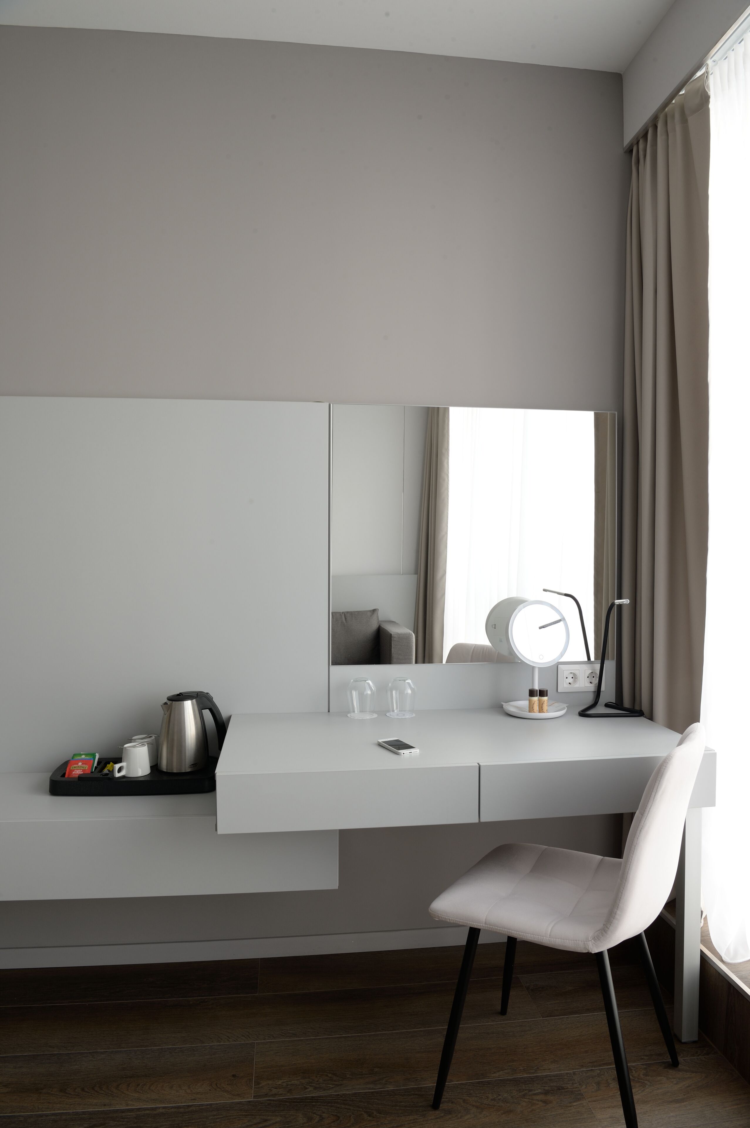 A modern, minimalist room with a white desk, a chair, a large mirror, and a bedside table with a lamp.