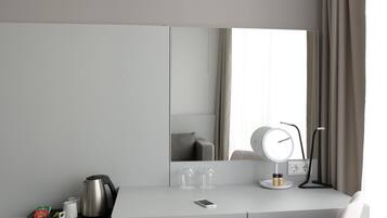 Una habitación moderna y minimalista con un escritorio blanco, una silla, un espejo grande y una mesita de noche con lámpara.