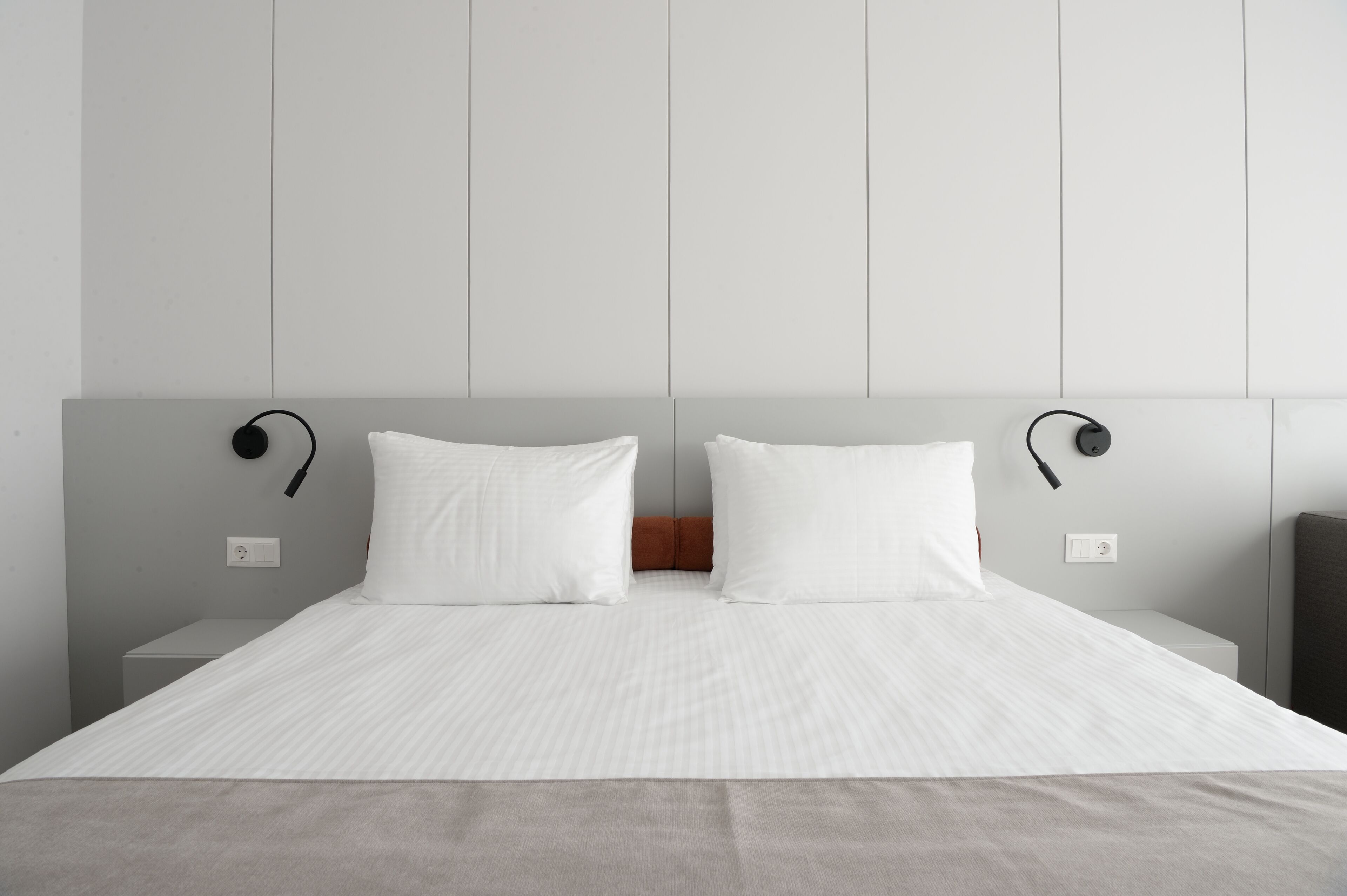 A neatly made bed with white linens, a grey headboard, and two bedside lamps.