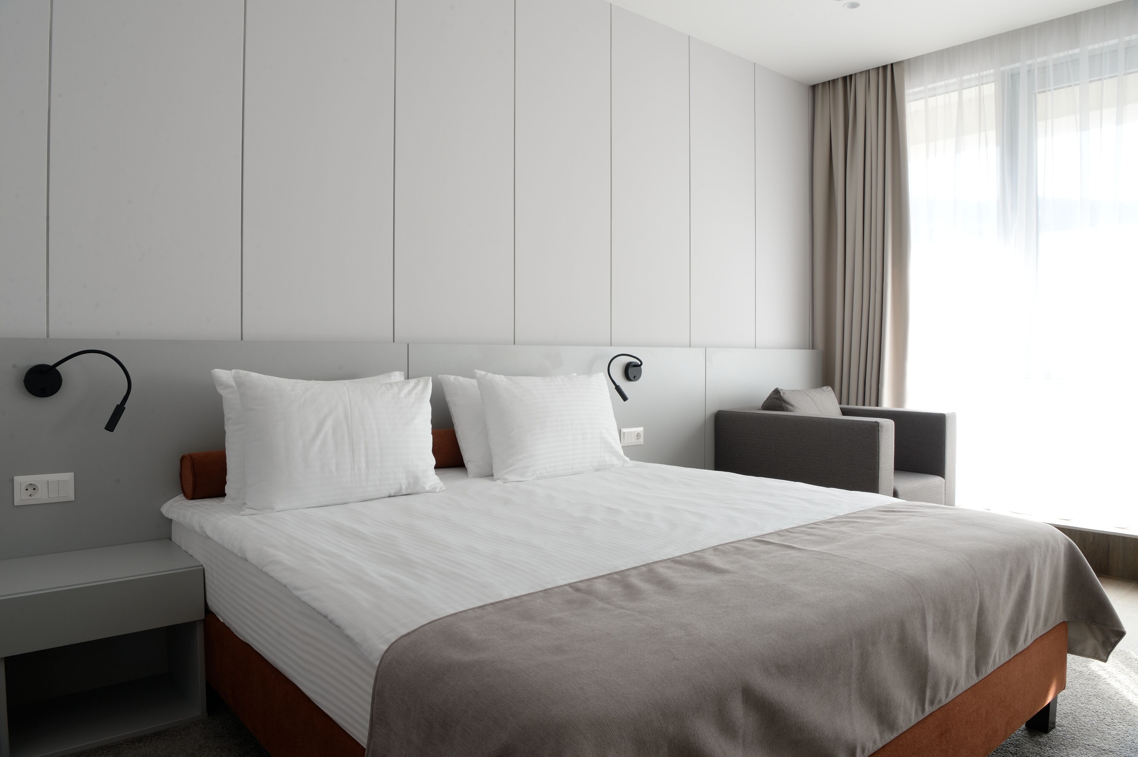 A modern hotel room with a large bed, a bedside table, a chair, and a large window with curtains.