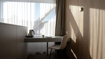 A hotel room with a wooden desk, a chair, a coffee maker, and a window with sheer curtains.