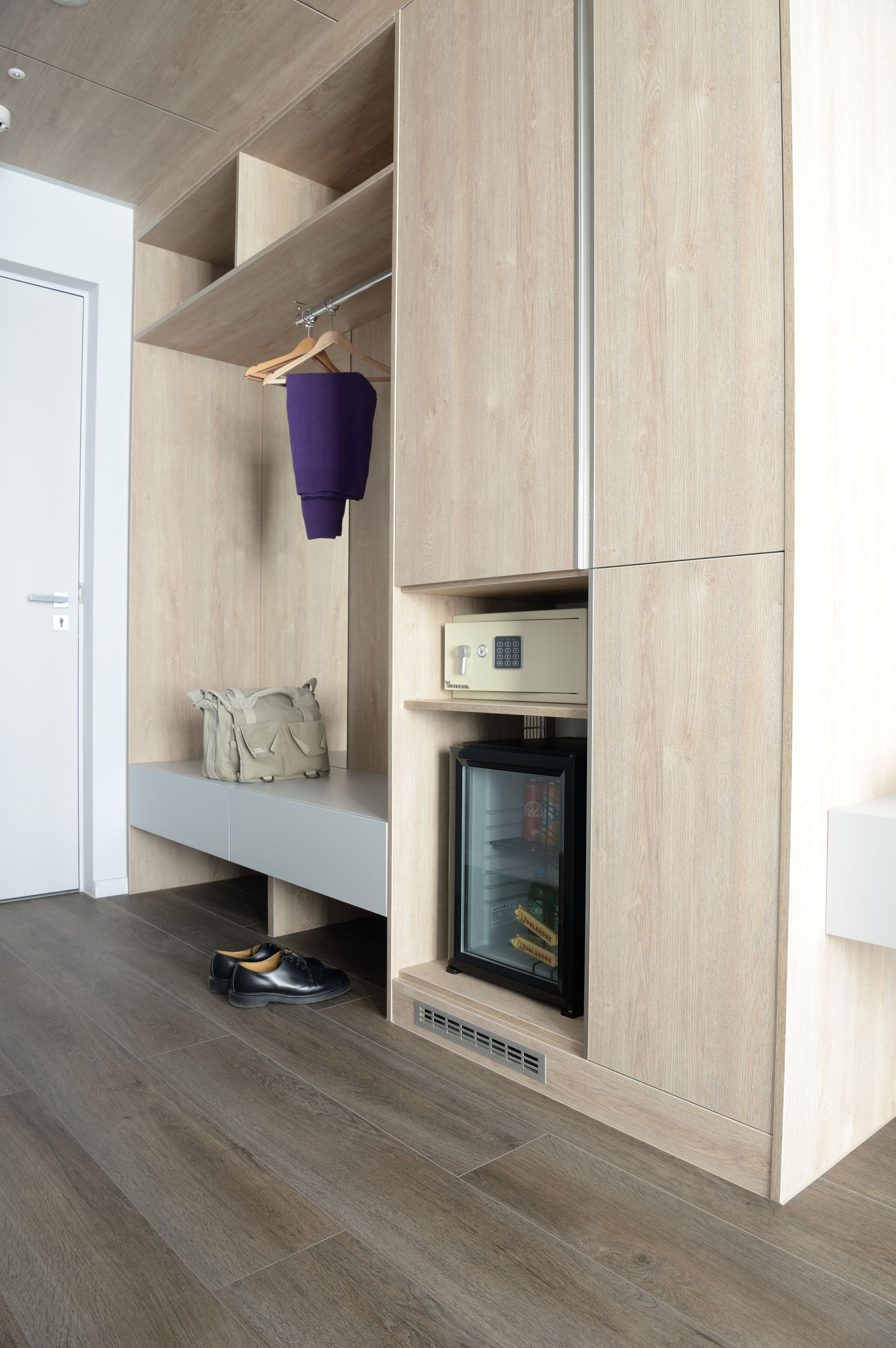 Een moderne kamer met een houten garderobe, een hangende kledingstuk, een klein plankje met een tas, en een vitrinekast met glazen deuren.