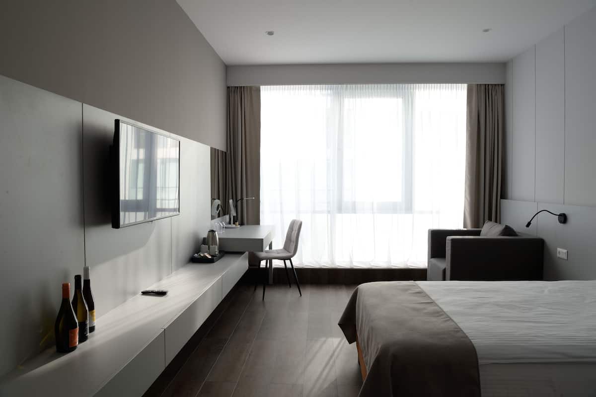A modern hotel room with a bed, a desk, a chair, a TV, and a window with curtains.