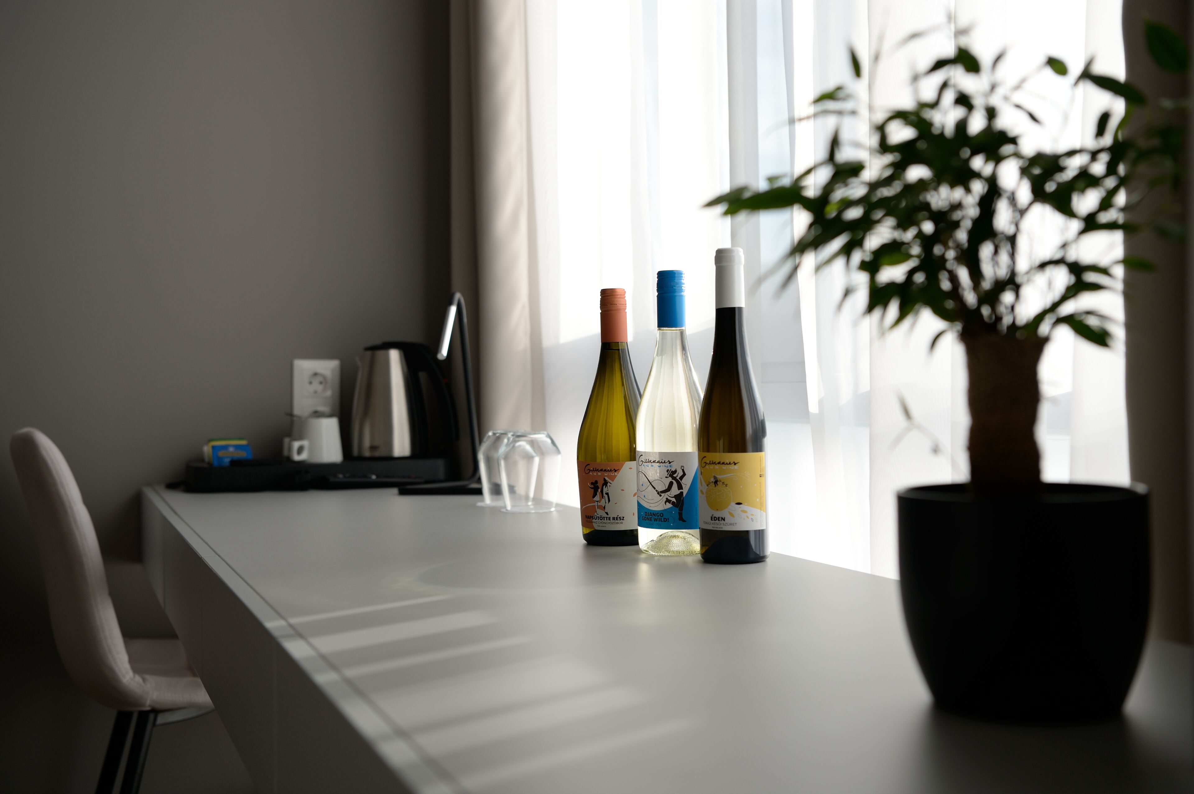 A table with three wine bottles, a coffee maker, and a potted plant in a room with a window.