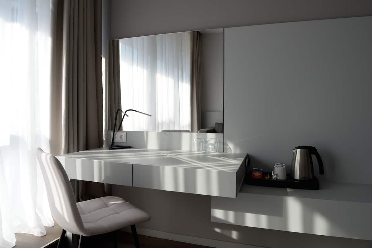 A modern hotel room with a built-in desk, a chair, a large mirror, and a coffee maker on a shelf.