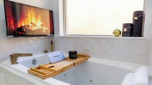 A modern bathroom with a bathtub, a wooden tray with a bottle and a candle, a TV displaying a fireplace, and a window with candles. - Modern Home with a Firepit, Heated Pool & Spa (Dallas)
