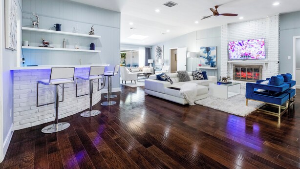 A modern living room with a white brick wall, a fireplace, a TV, and a ceiling fan. - Modern Home with a Firepit, Heated Pool & Spa (Dallas)