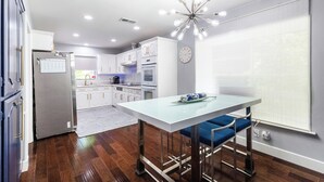A modern kitchen with white cabinets, a stainless steel refrigerator, and a dining area with a glass table and blue chairs. - Modern Home with a Firepit, Heated Pool & Spa (Dallas)