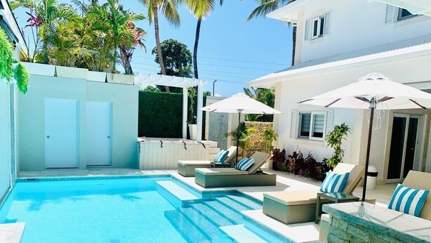 A swimming pool with lounge chairs and umbrellas, surrounded by a white building with a stone wall and plants.