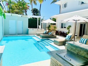 A swimming pool with lounge chairs and umbrellas, surrounded by a white building with a stone wall and plants. - Villa Bella Vida (Cabarete)