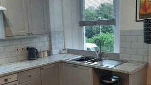 Eine Küche mit weißen Schränken, einem Spülbecken, einem Fenster mit Jalousien und einem Mülleimer.