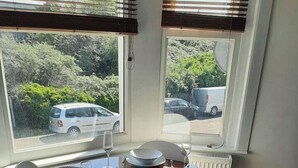 Ein Esstisch für vier Personen, mit weißen Stühlen, befindet sich in der Nähe eines Fensters mit Jalousien und bietet Blick auf einen Parkplatz und Grünflächen.