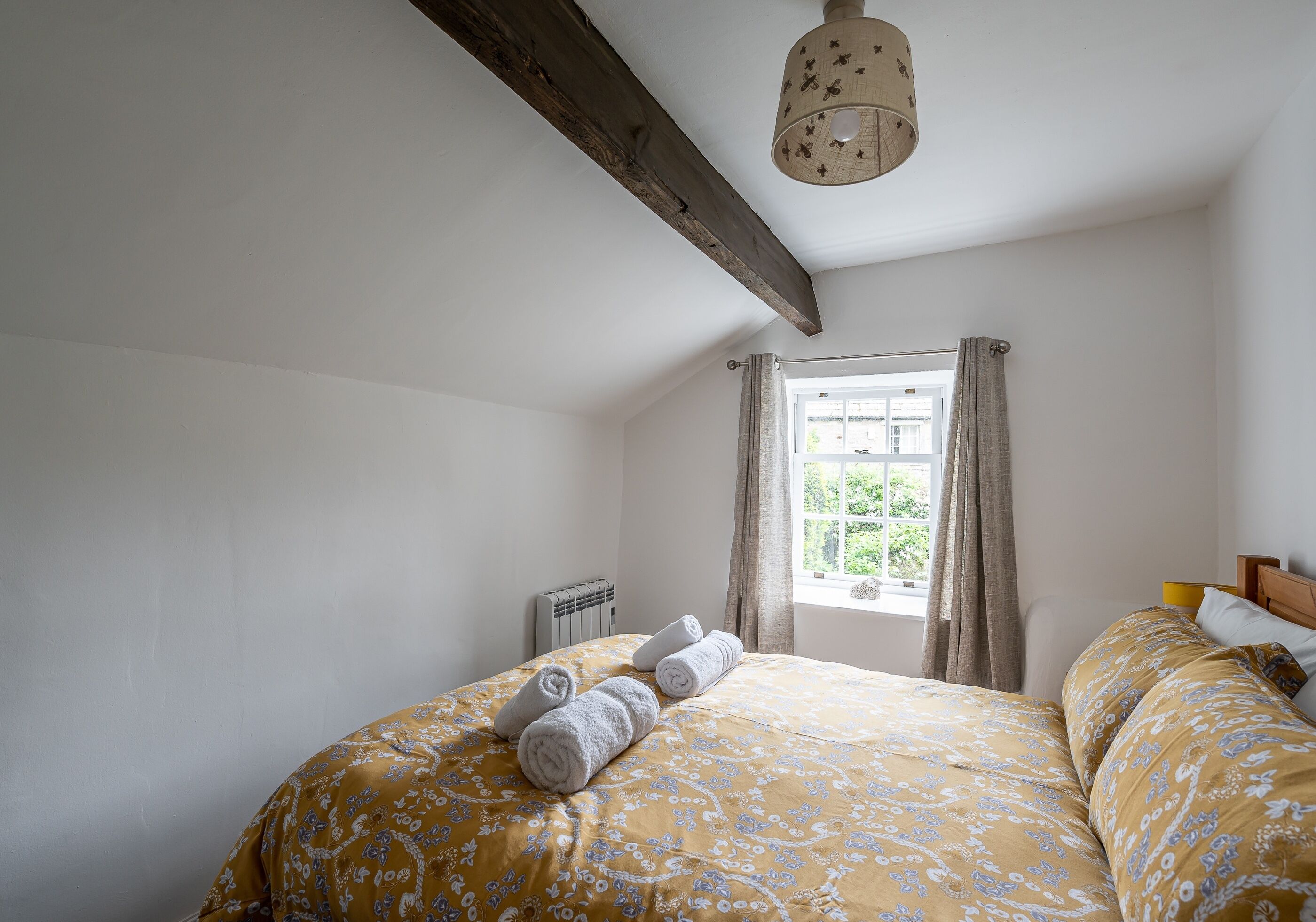 A bedroom with a bed, a window with curtains, a radiator, and a wooden ceiling beam.