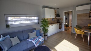 A modern living space with a grey sofa, a dining table with yellow chairs, a kitchen area, and a large window providing natural light.