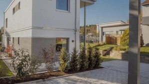 A modern two-story building with a flat roof, a garage, and a small garden area with a patio and potted plants.