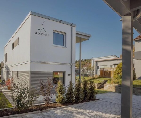 Um edifício moderno de dois andares com telhado plano, garagem e um pequeno jardim com pátio e plantas em vasos.