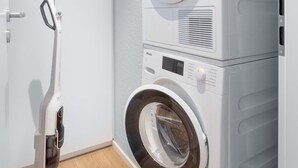 A compact laundry area with a stacked washer and dryer, a vacuum cleaner, and a small window.