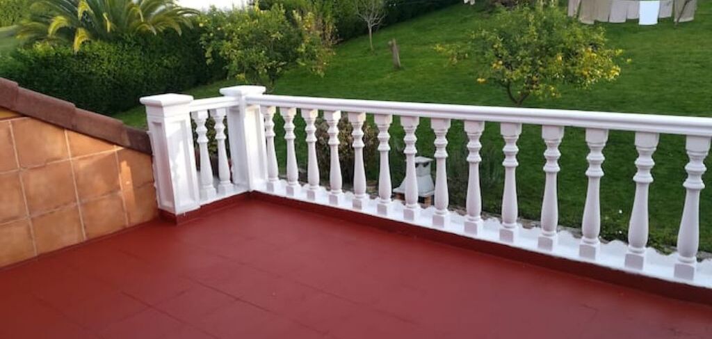 Un balcón con piso rojo y barandilla blanca, con vista a un césped y jardín bien cuidados.