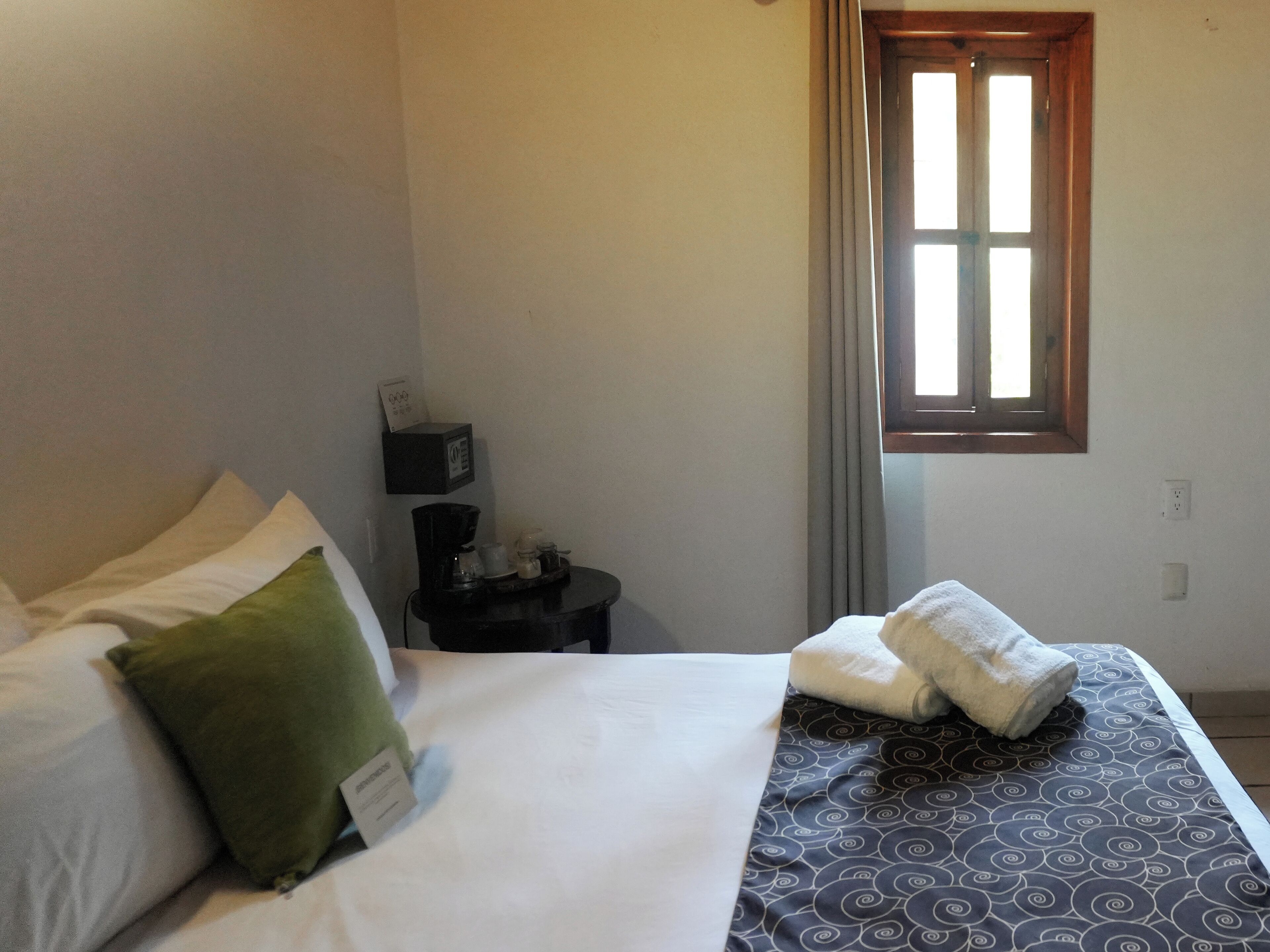 A neatly made bed with pillows and a blanket, a small bedside table with a lamp, a window with curtains, and a wall-mounted power outlet.