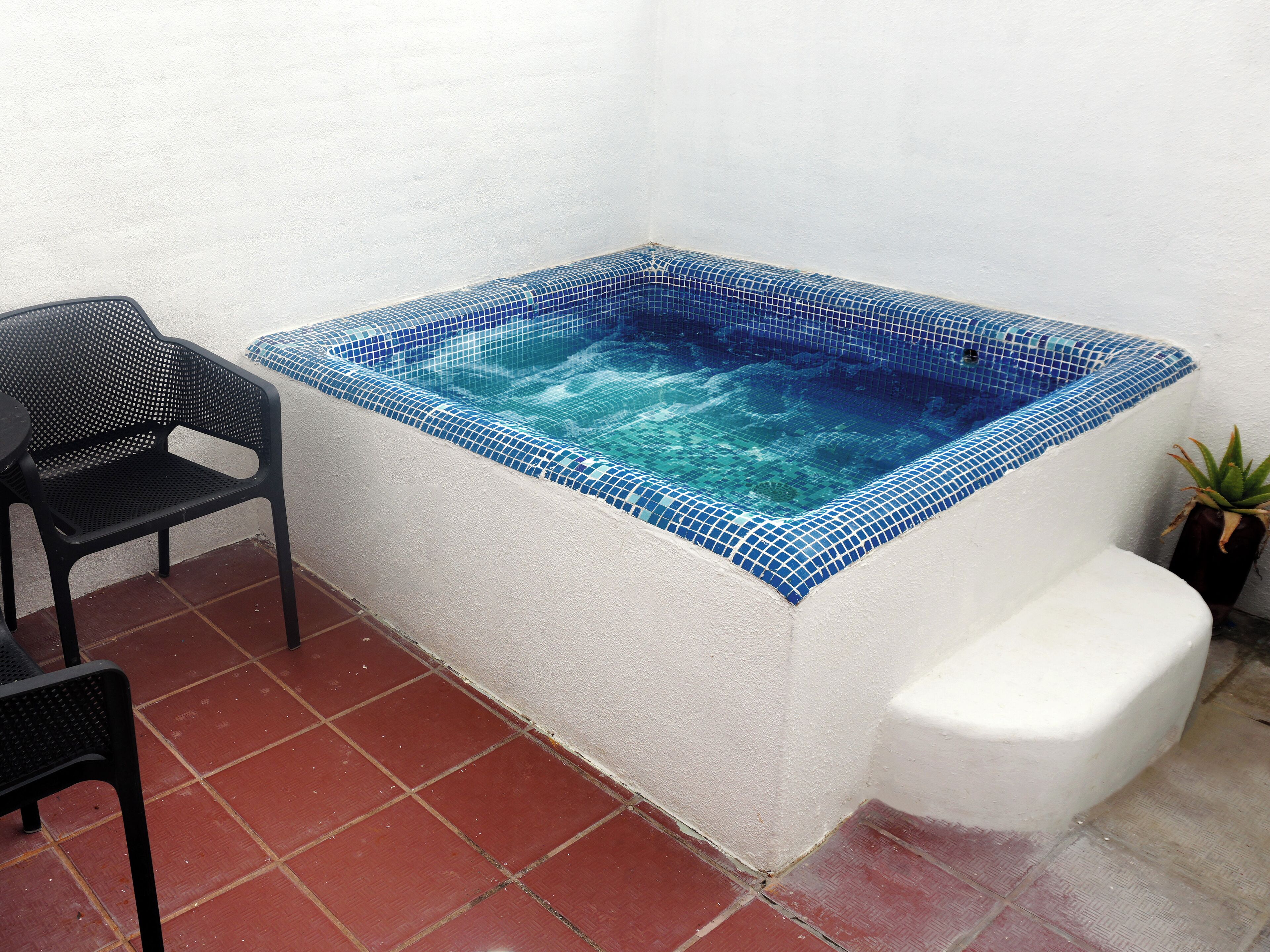 Un bain à remous rectangulaire, orné de mosaïques bleues, avec une chaise en métal noir et une plante en pot placée à côté.