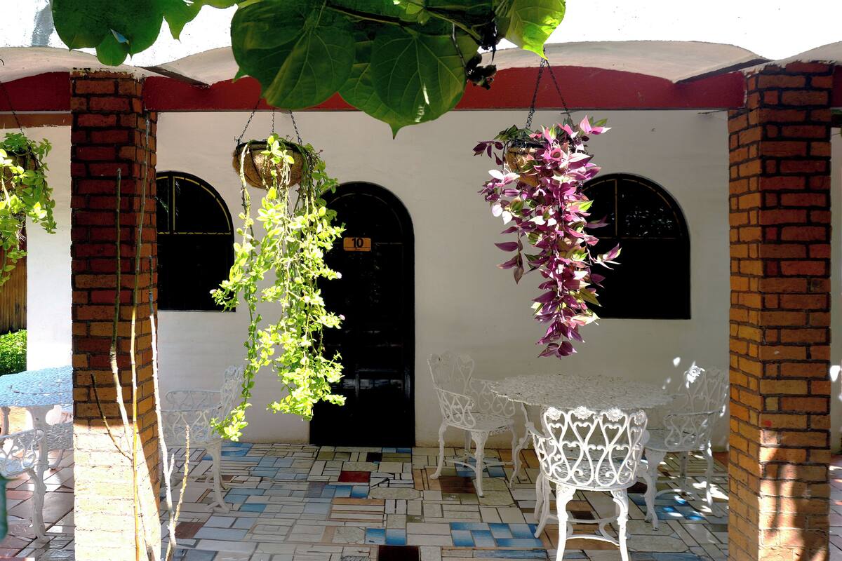 A courtyard with a tiled floor, white chairs, hanging plants, and a brick wall with two black doors.