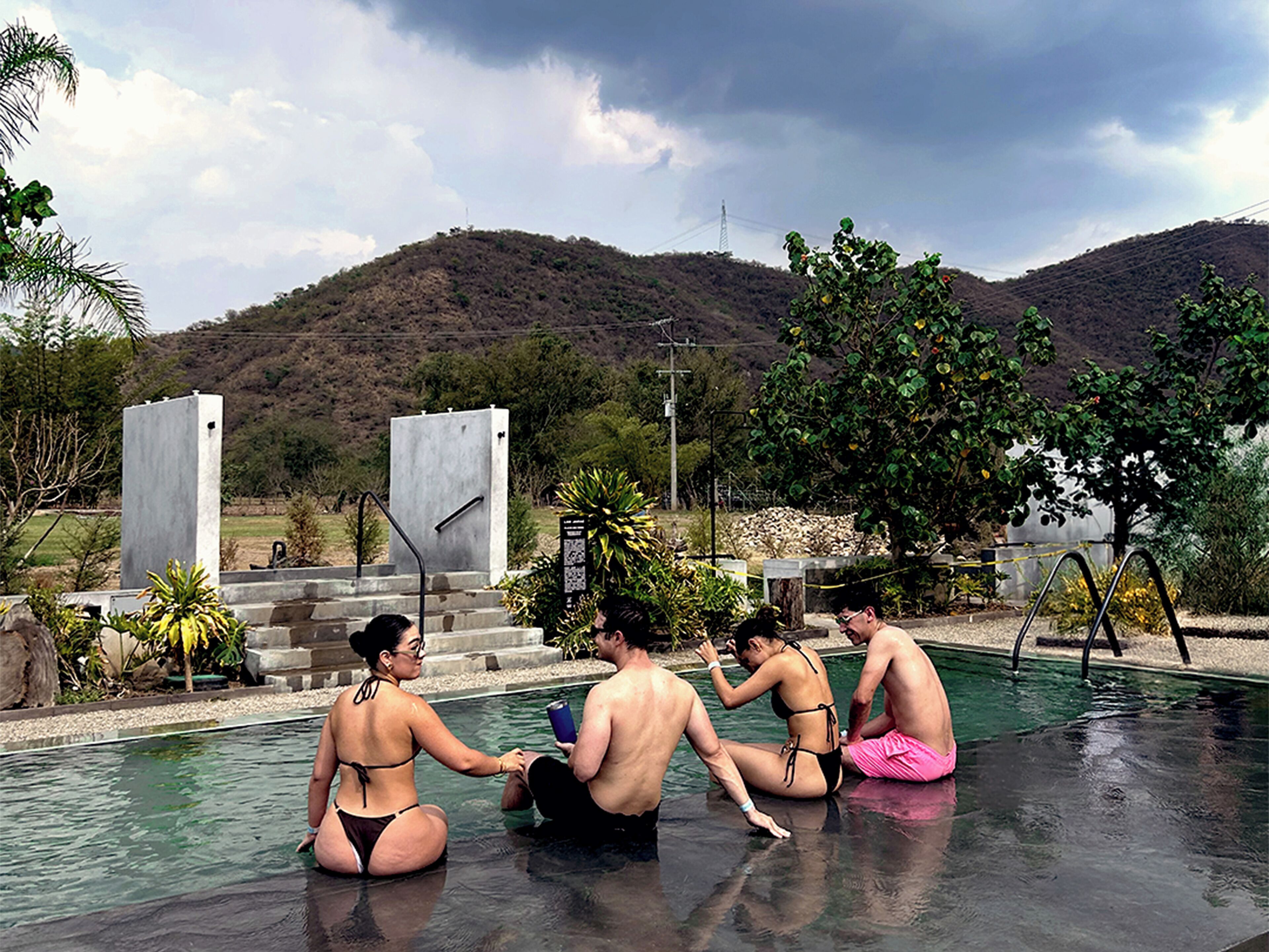 A swimming pool with people relaxing, surrounded by hills and a cloudy sky.