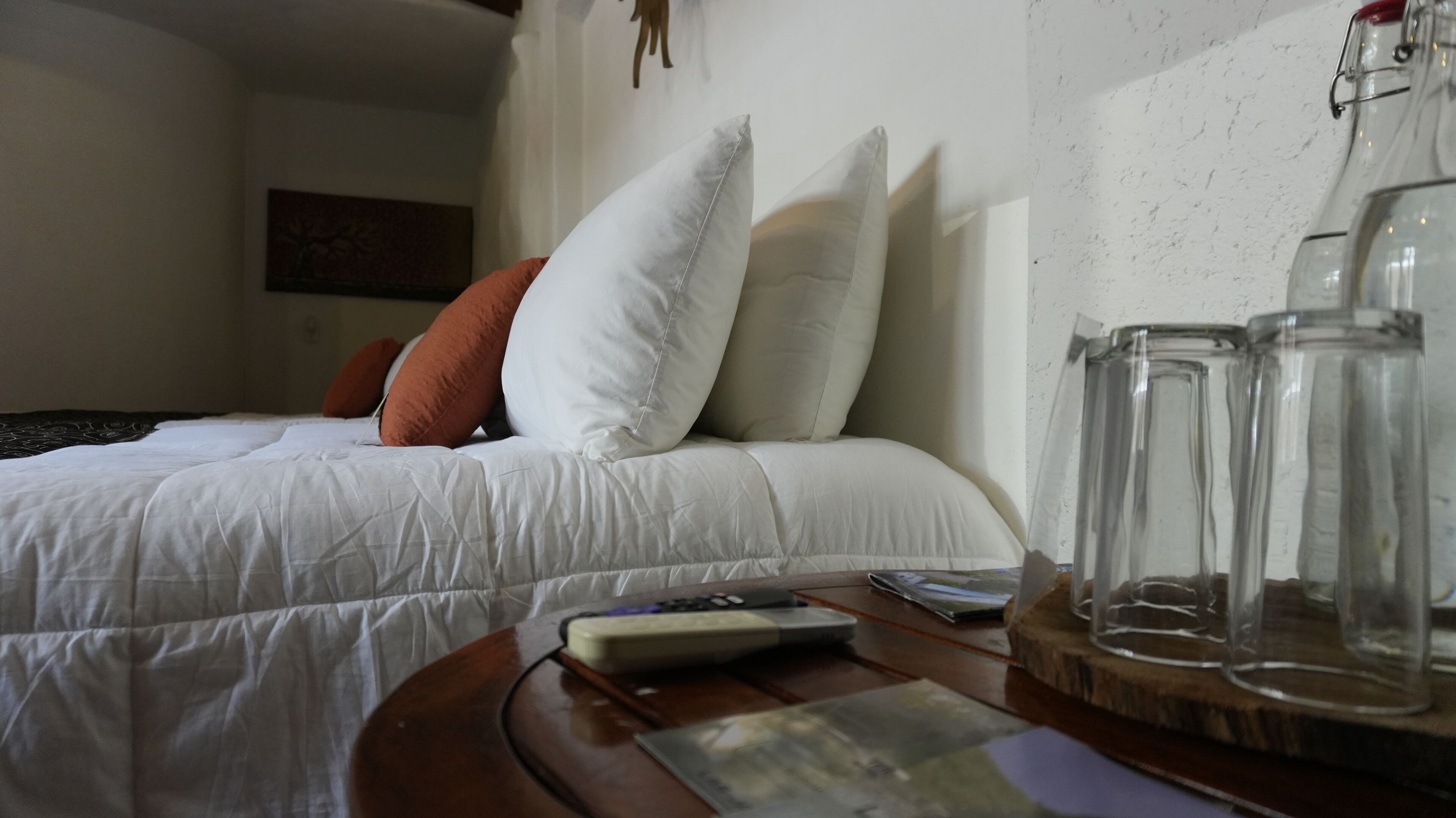 Una cama bien tendida con almohadas blancas y anaranjadas, un control remoto y un tarro de vidrio sobre una mesita de madera.