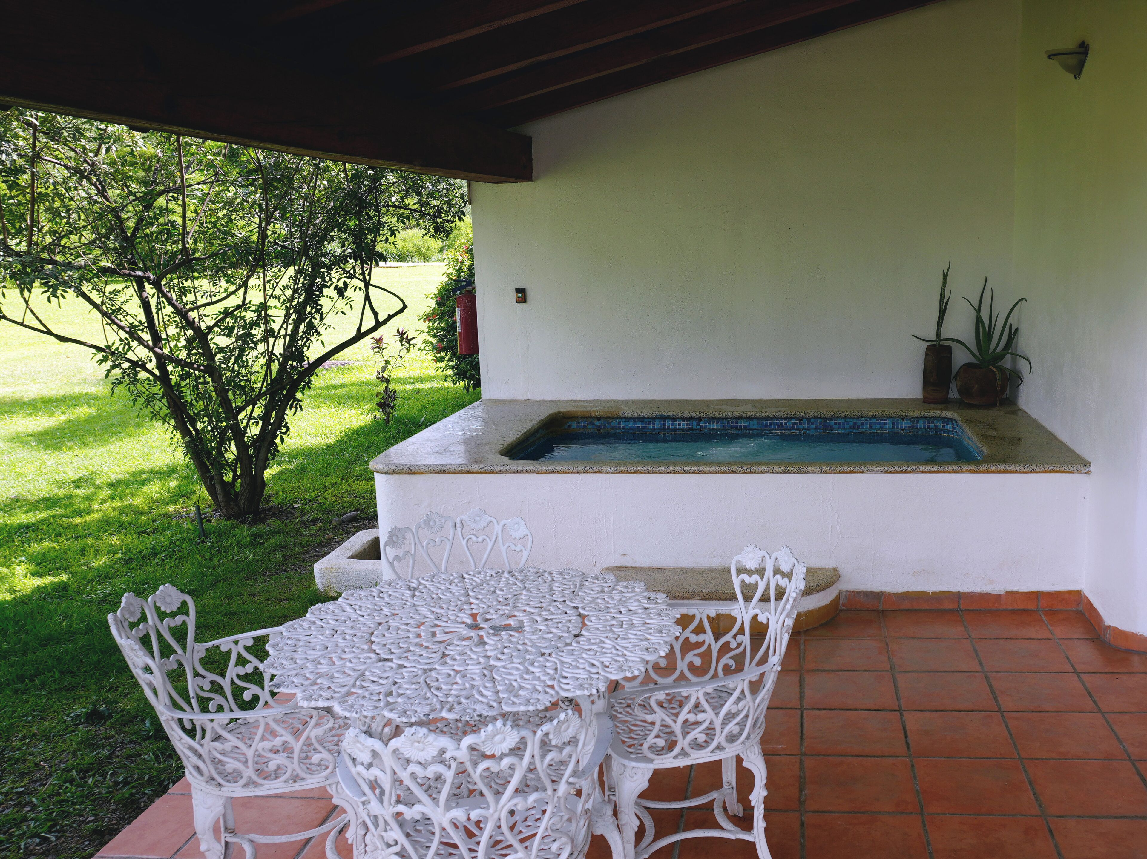 Ein überdachter Patio-Bereich mit Pool, weißer Gartenmöbel und Blick auf einen Garten.