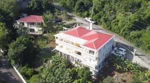 Une maison à deux étages, avec un toit rouge, entourée d’une végétation luxuriante.