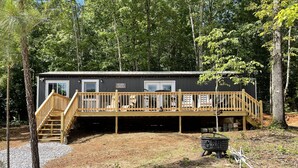Une cabane dotée d’une terrasse en bois, d’un petit espace extérieur aménagé pour s’asseoir et d’un chemin de gravier menant à l’entrée.