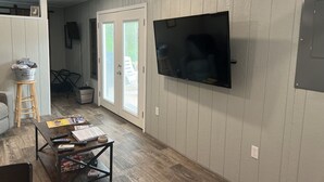 Un salon doté d’un téléviseur à écran plat fixé au mur, d’une table basse avec des télécommandes, et d’une porte donnant sur l’extérieur.