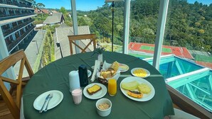 A table set with breakfast in a covered outdoor area with a view of a sports field and forested hills. - Apt Quatre Saisons (Campos do Jordão)