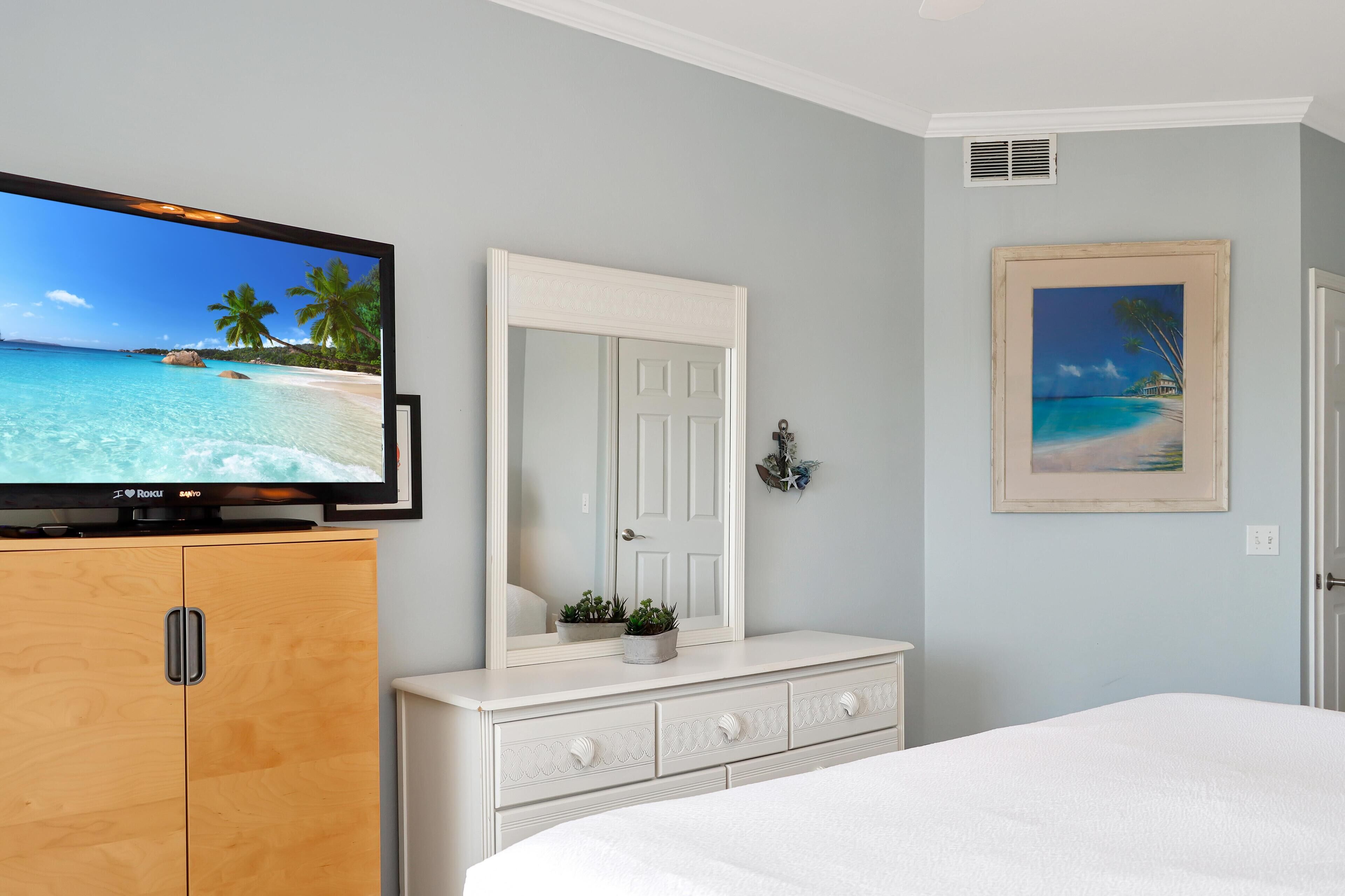 A bedroom with a bed, a dresser, a TV, a mirror, and a framed painting.