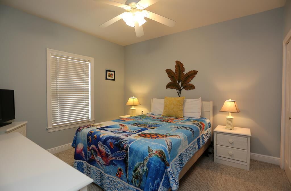 A bedroom with a bed, two bedside tables with lamps, a ceiling fan, a window with blinds, and a television on a stand.