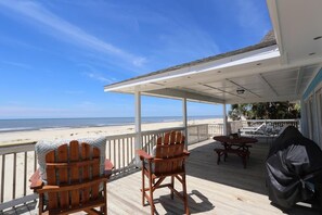 Una terraza de madera con dos sillas Adirondack, una mesa de picnic y una parrilla, con vista a una playa.