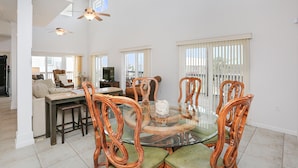 A spacious dining area with a glass table and wooden chairs, a kitchen in the background, and a balcony with white blinds. - Beach Please (Port St. Joe)
