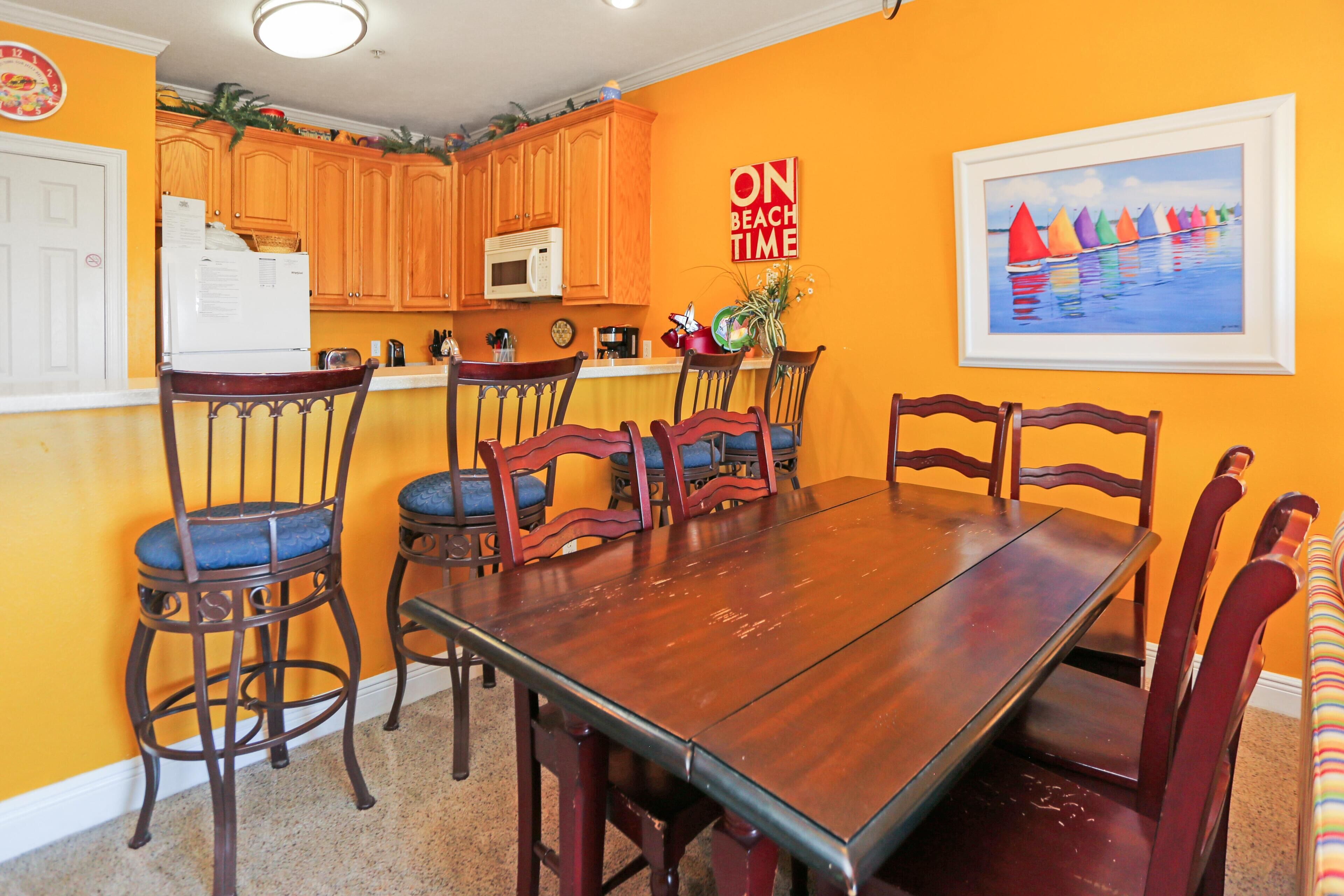 A dining area with a wooden table and chairs, a kitchen with wooden cabinets, a microwave, and a painting on the wall.