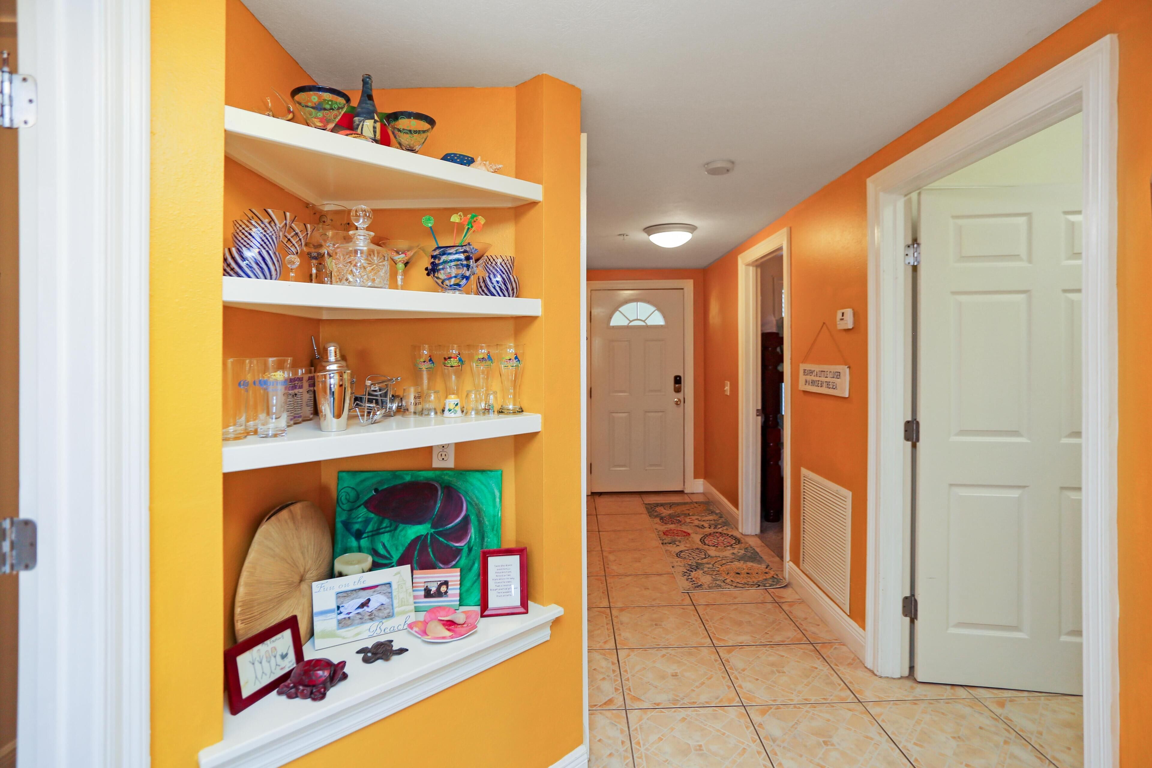 Un couloir aux murs orange, avec des étagères blanches où sont exposés des objets en verre et des éléments de décoration, et une porte blanche au fond.