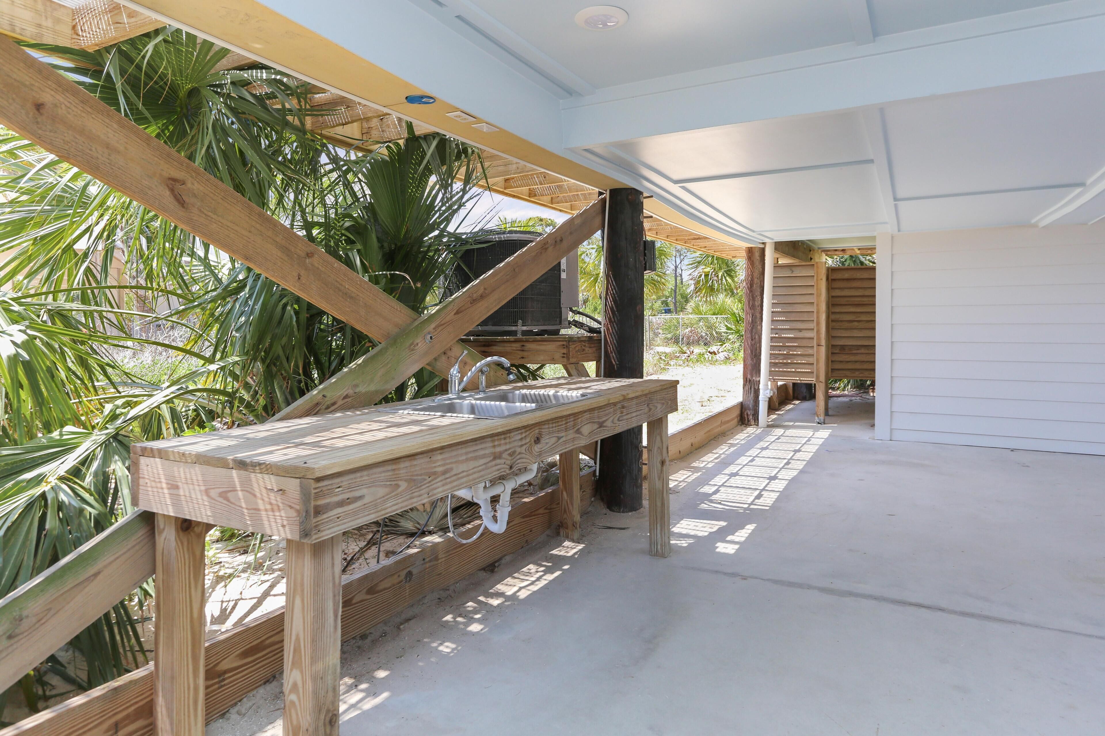 Un espace cuisine extérieur avec une table en bois, un évier et un barbecue, entouré de palmiers et avec un bâtiment blanc en arrière-plan.