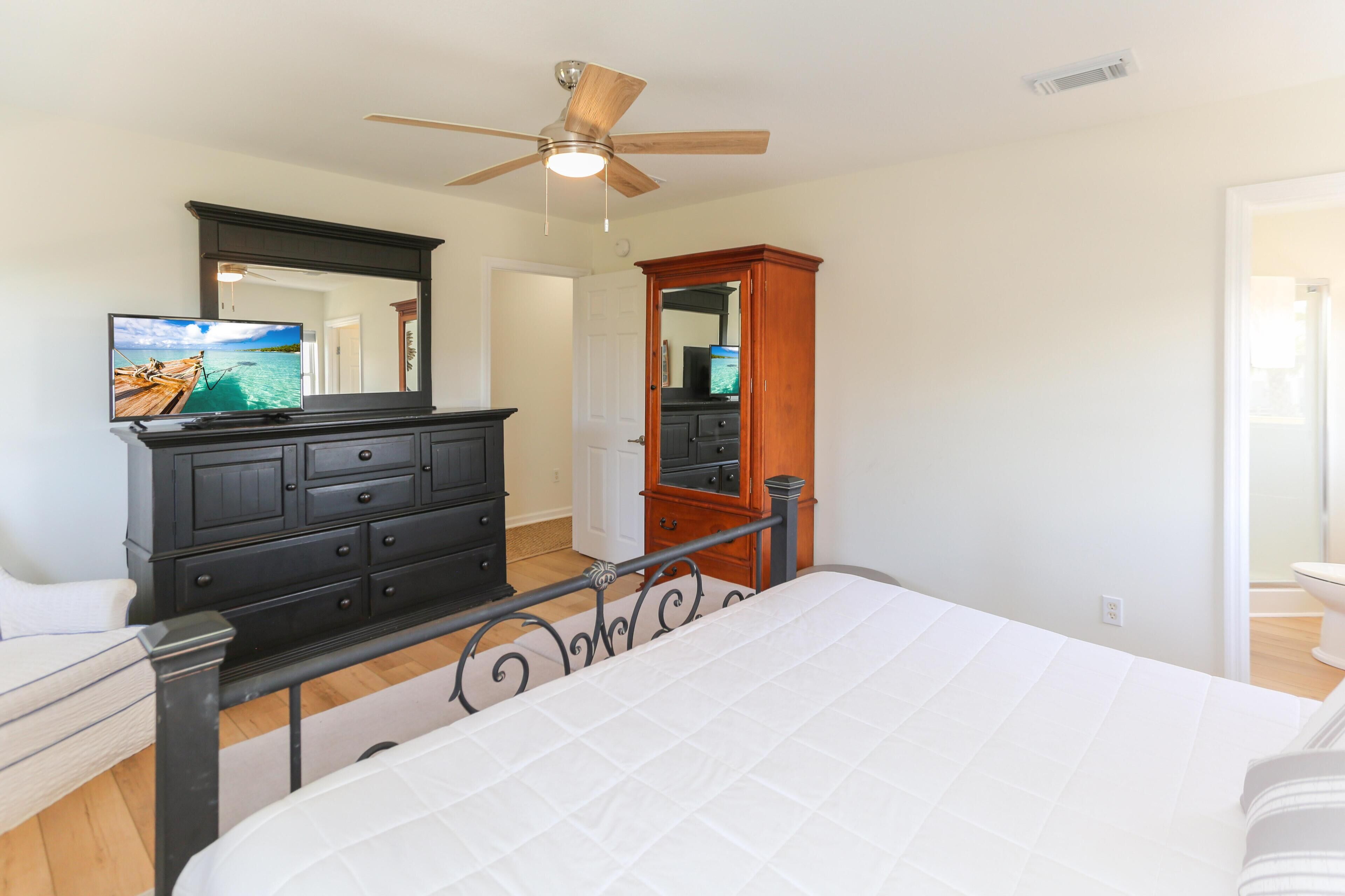 A bedroom with a bed, a dresser, a television, a ceiling fan, and a door leading to another room.