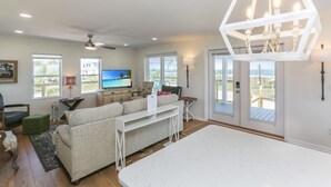 A spacious living room with a large sofa, a white coffee table, and a television. There are multiple windows providing natural light and views of the outdoors. - Brodie House (Port St. Joe)
