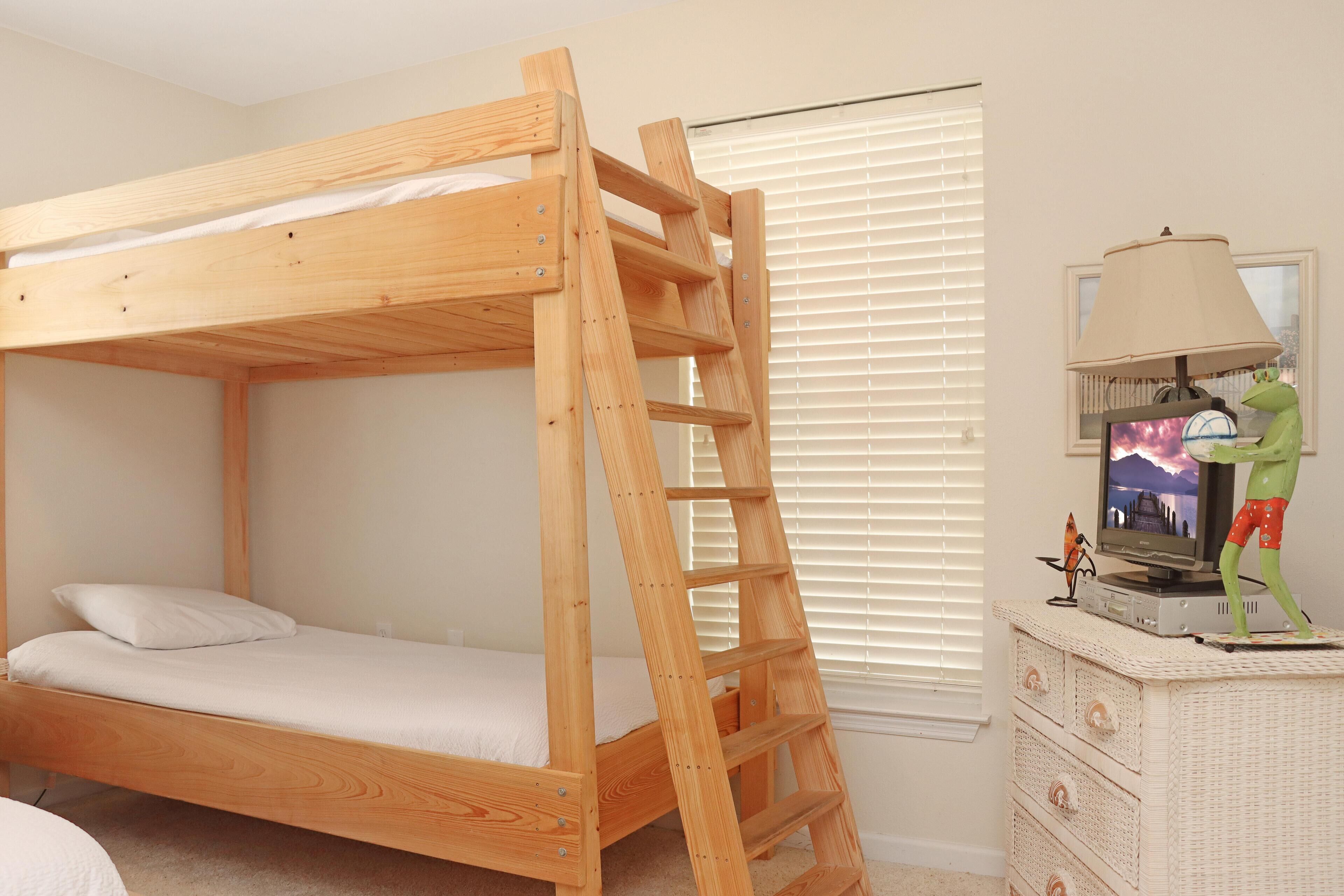 A bunk bed with a wooden ladder, a television on a dresser, and a window with blinds.