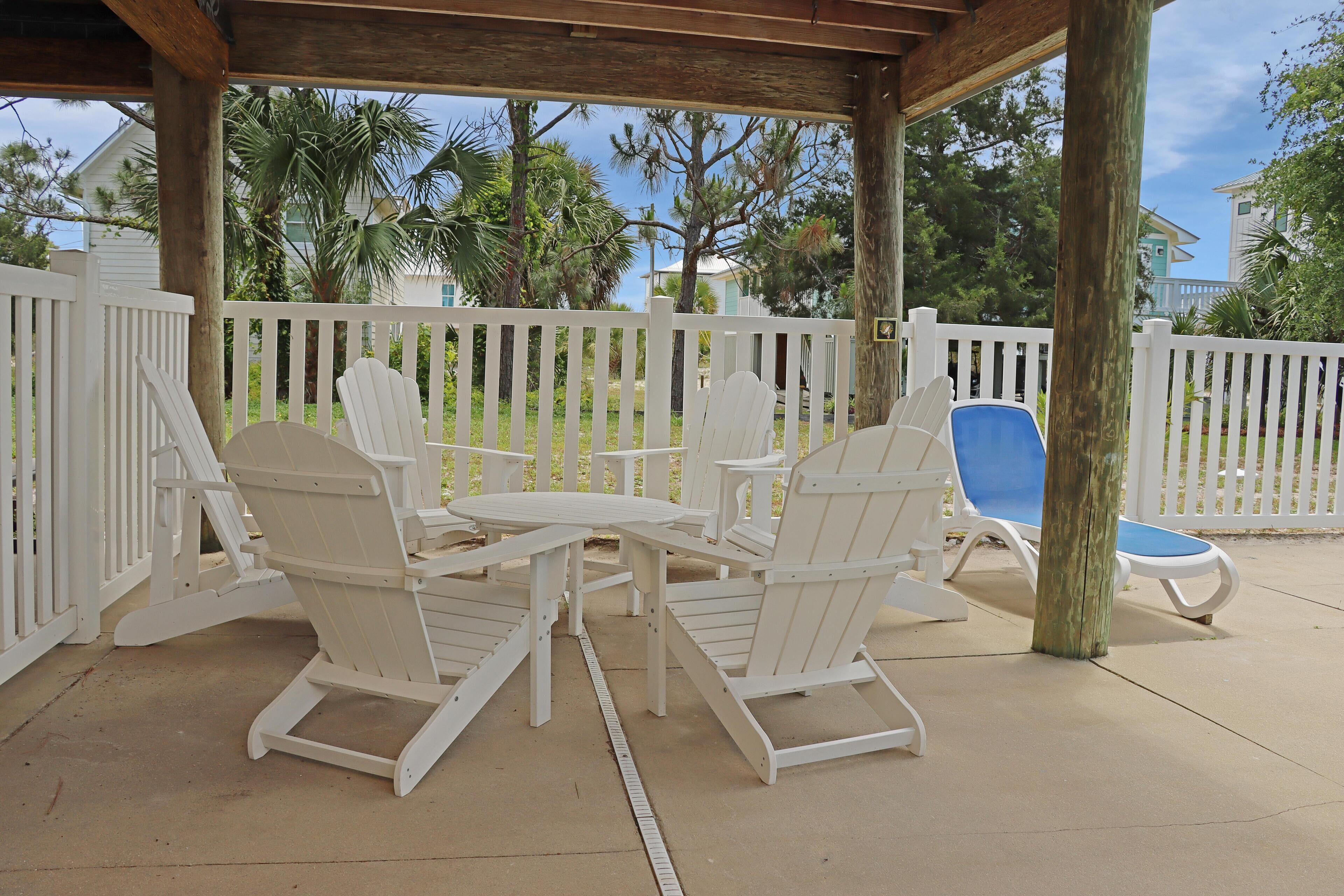 A covered patio area with white Adirondack chairs and a table, surrounded by a white picket fence.