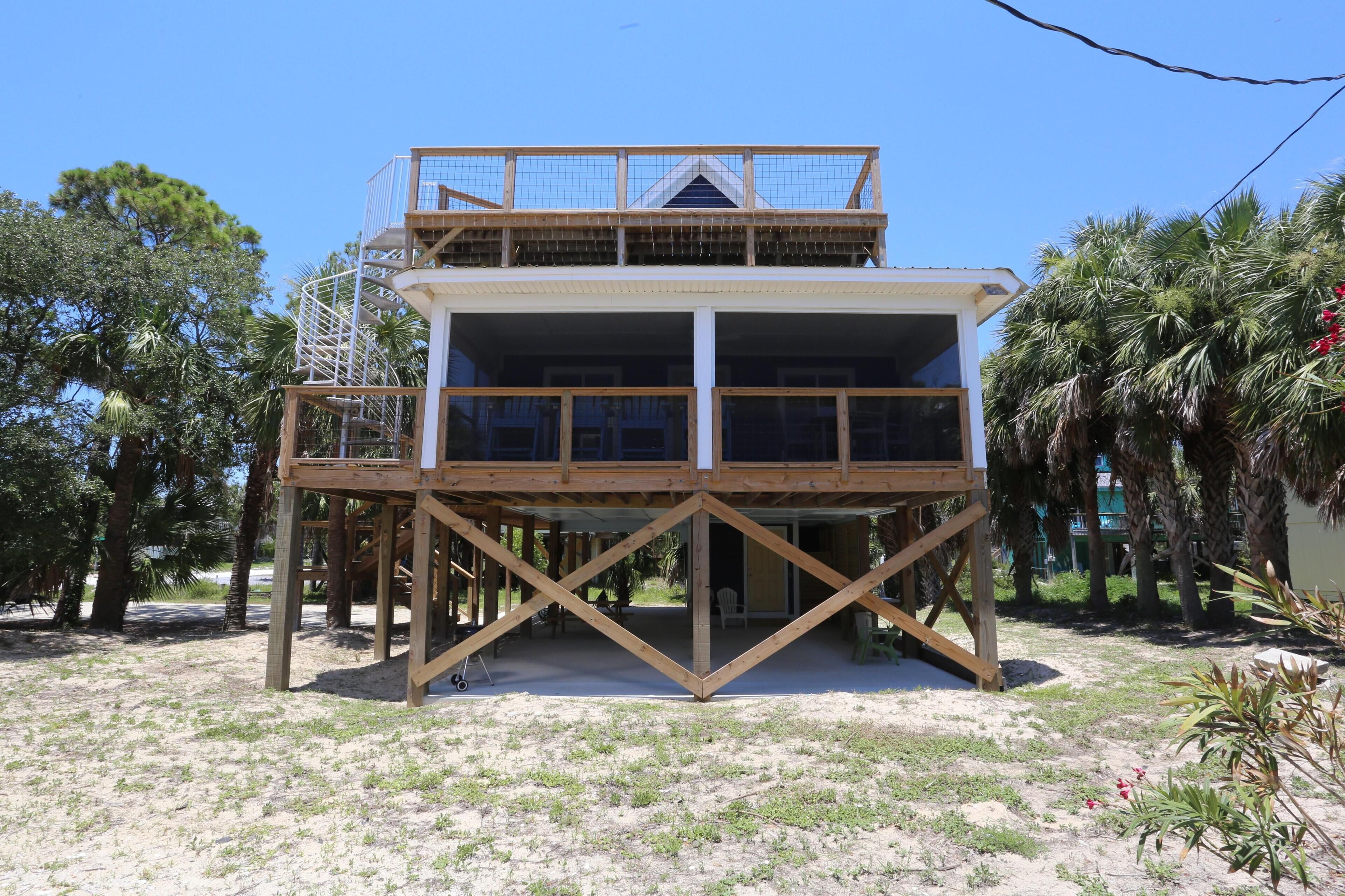 A two-story wooden structure with a balcony, surrounded by palm trees and a sandy ground.