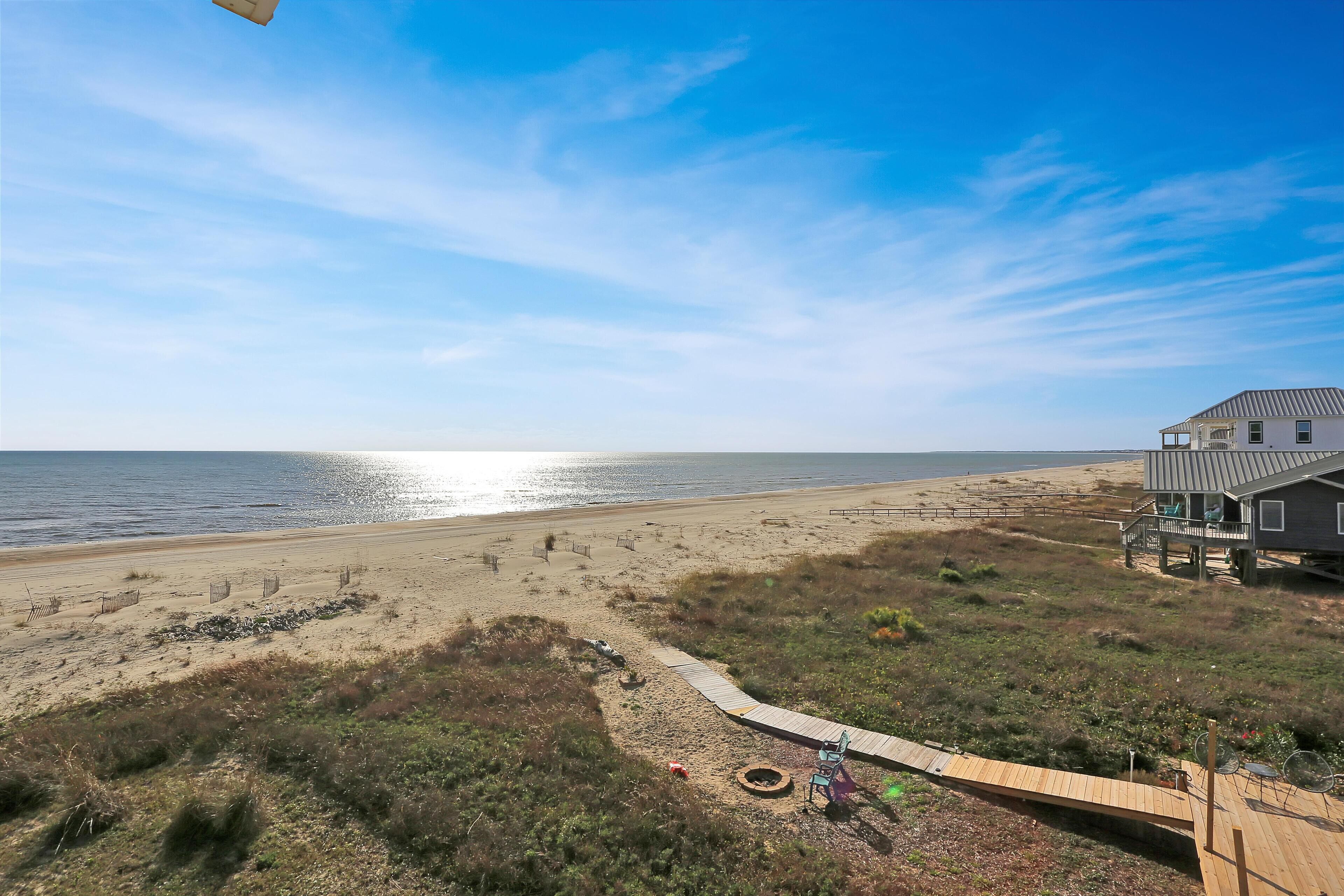 A beach house with a wooden deck, a paved path leading to the beach, and a view of the ocean.