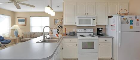 A kitchen with white cabinets, a microwave, stove, and refrigerator. There is a ceiling fan and a window with blinds.