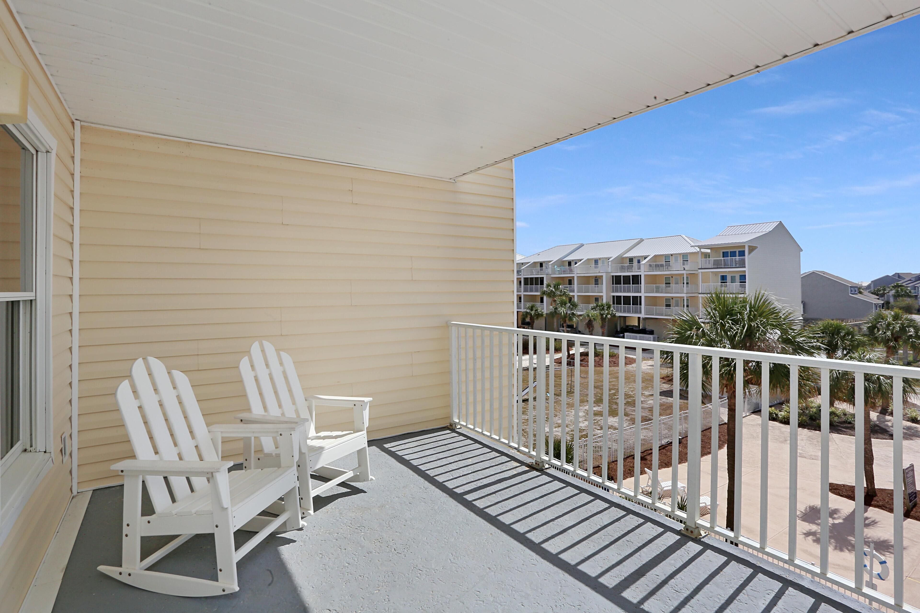 A balcony with two rocking chairs and a small table, overlooking a landscaped area with palm trees and a building in the background.