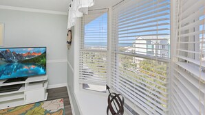 Quarto com televisão em suporte branco, janela com persianas e vista para um edifício.