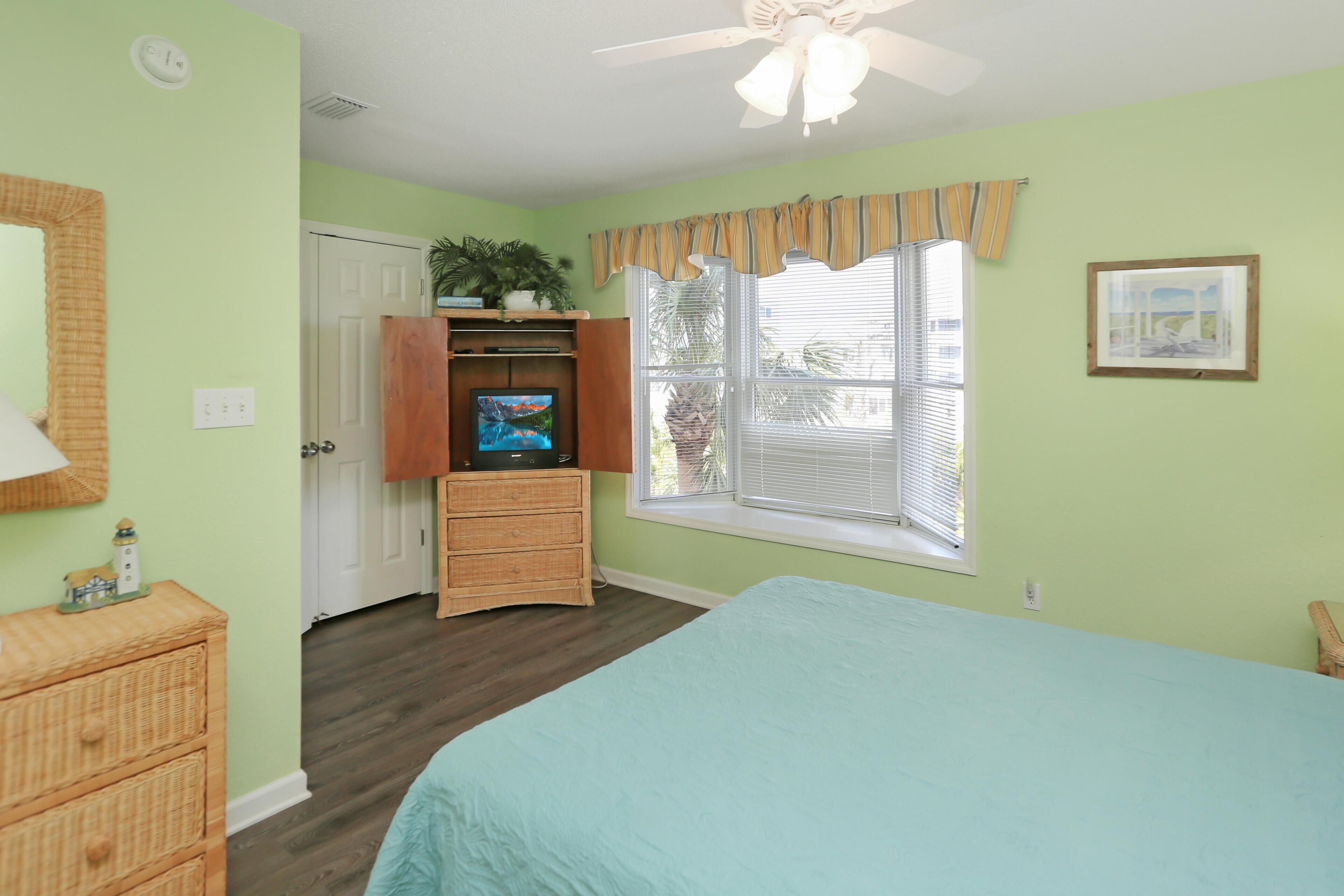 A bedroom with a bed, a television on a stand, a window with blinds, a ceiling fan, and a door.