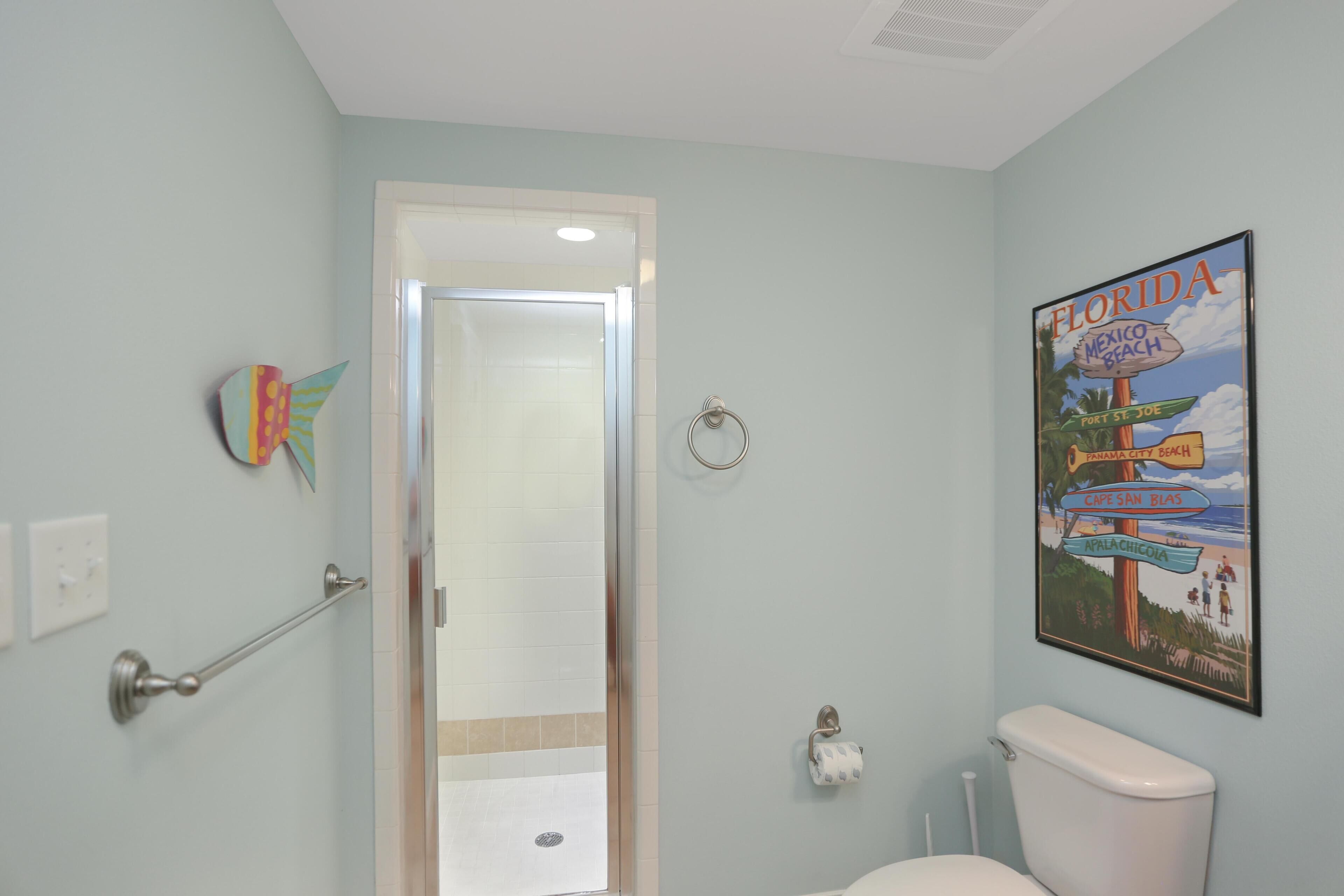 A bathroom with a toilet, a shower, a towel rack, a toilet paper holder, and a Florida beach sign.